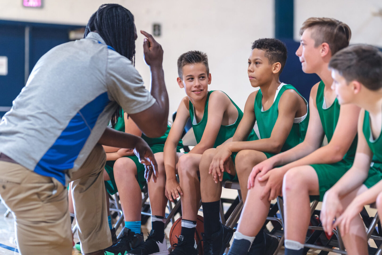 Several players sit on the sideline seats during the game with their coach and wait for their chance to get in and play. He's looking at one and trying to get his attention while the player is grinning at one of his teammates.